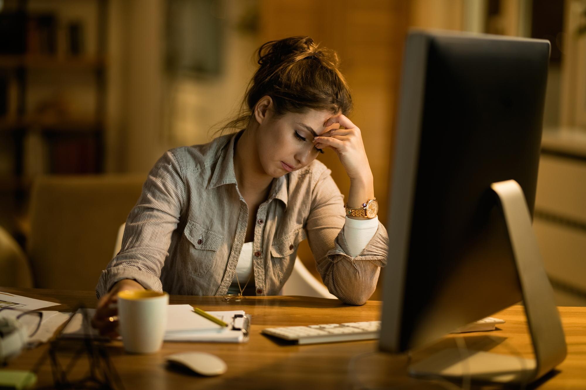 Tired employee resting her head in her hand at a desk in the evening, an early visual sign of workplace burnout
