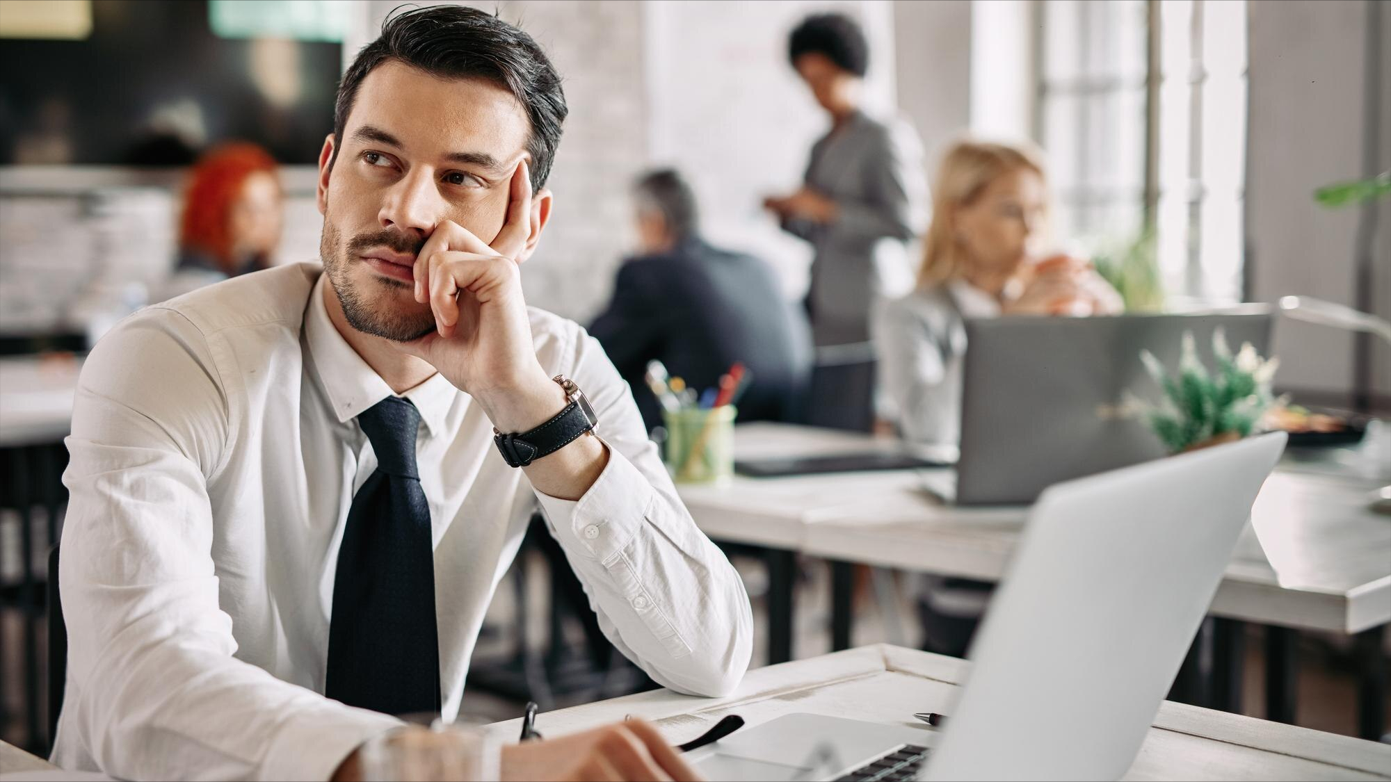 Pensive manager sitting at a desk in an office, reflecting on why employee appreciation programs fail while colleagues work in the background